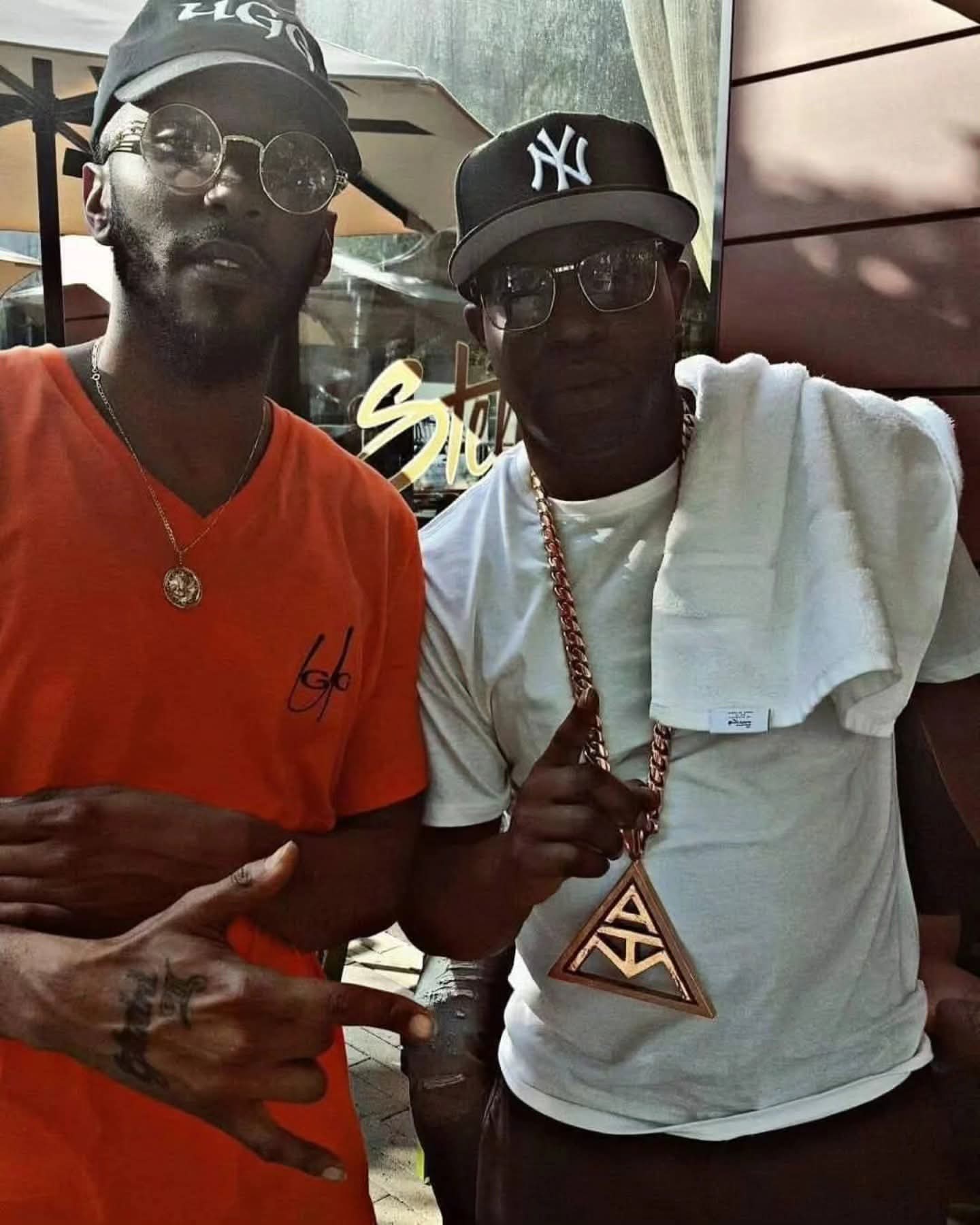Two men posing, one in orange and another wearing a large gold triangle pendant.