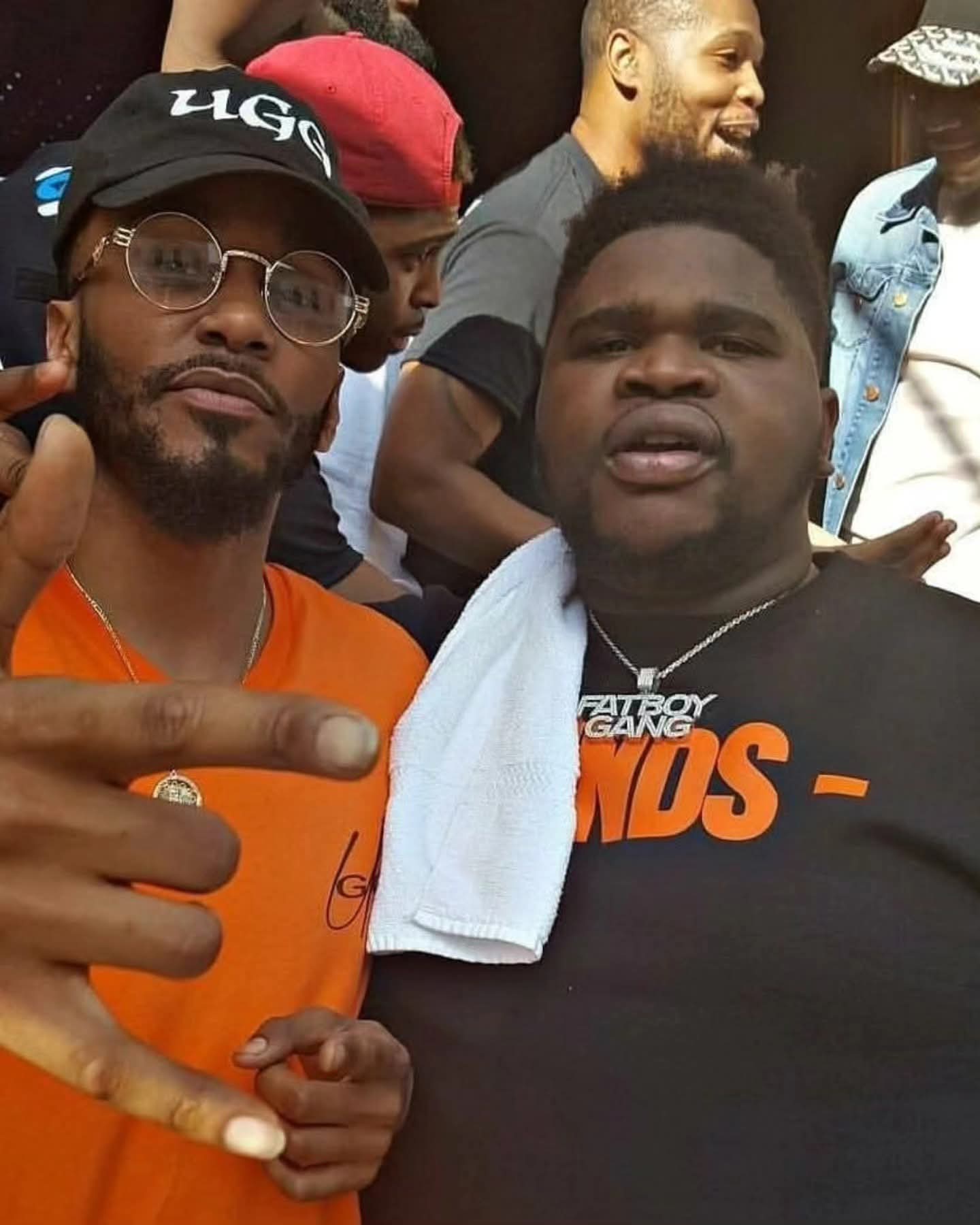 Two men posing; one wears an orange shirt and glasses, the other a black tee.