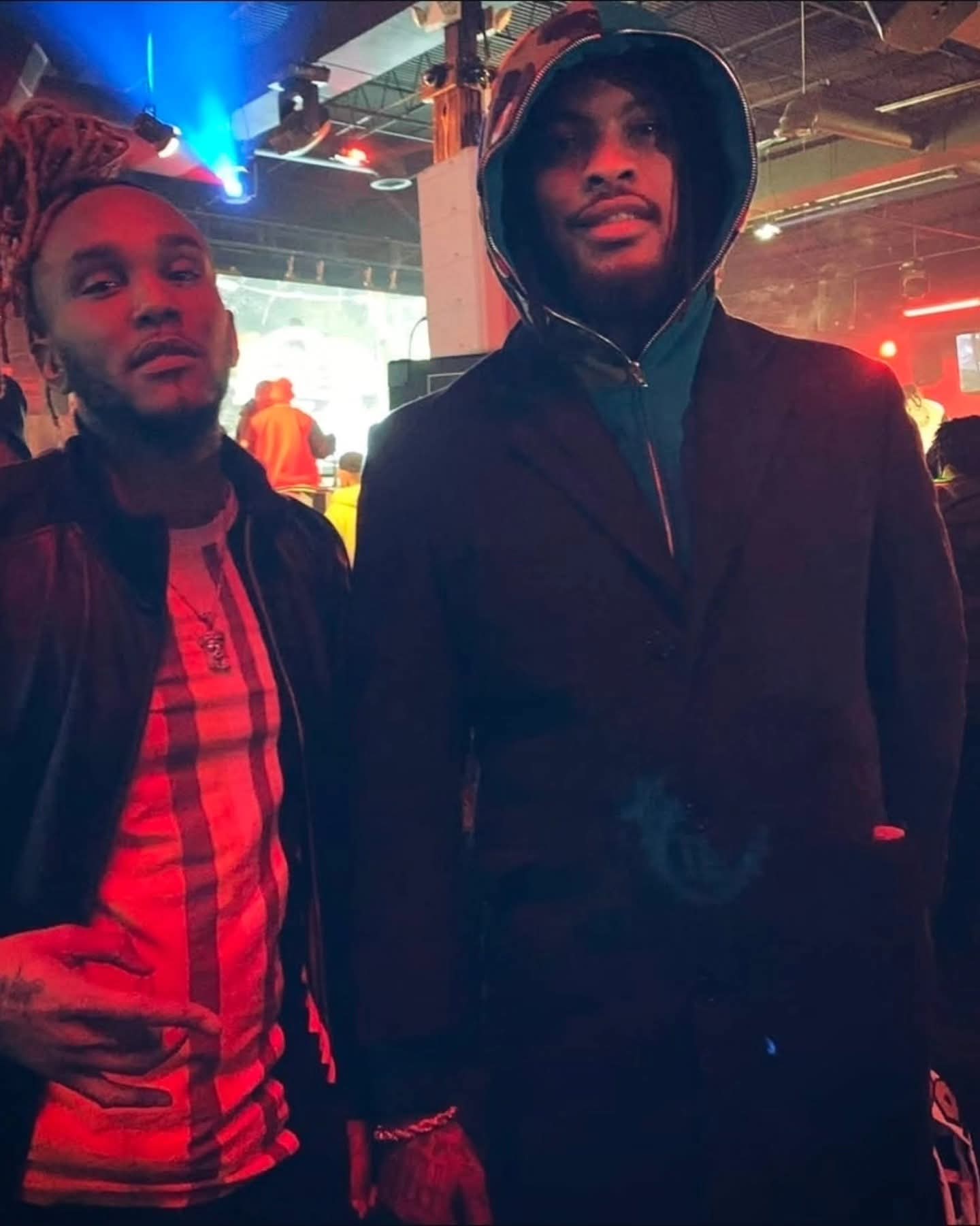 Waka Flocka Flame and a man pose in a club with red and blue lighting.