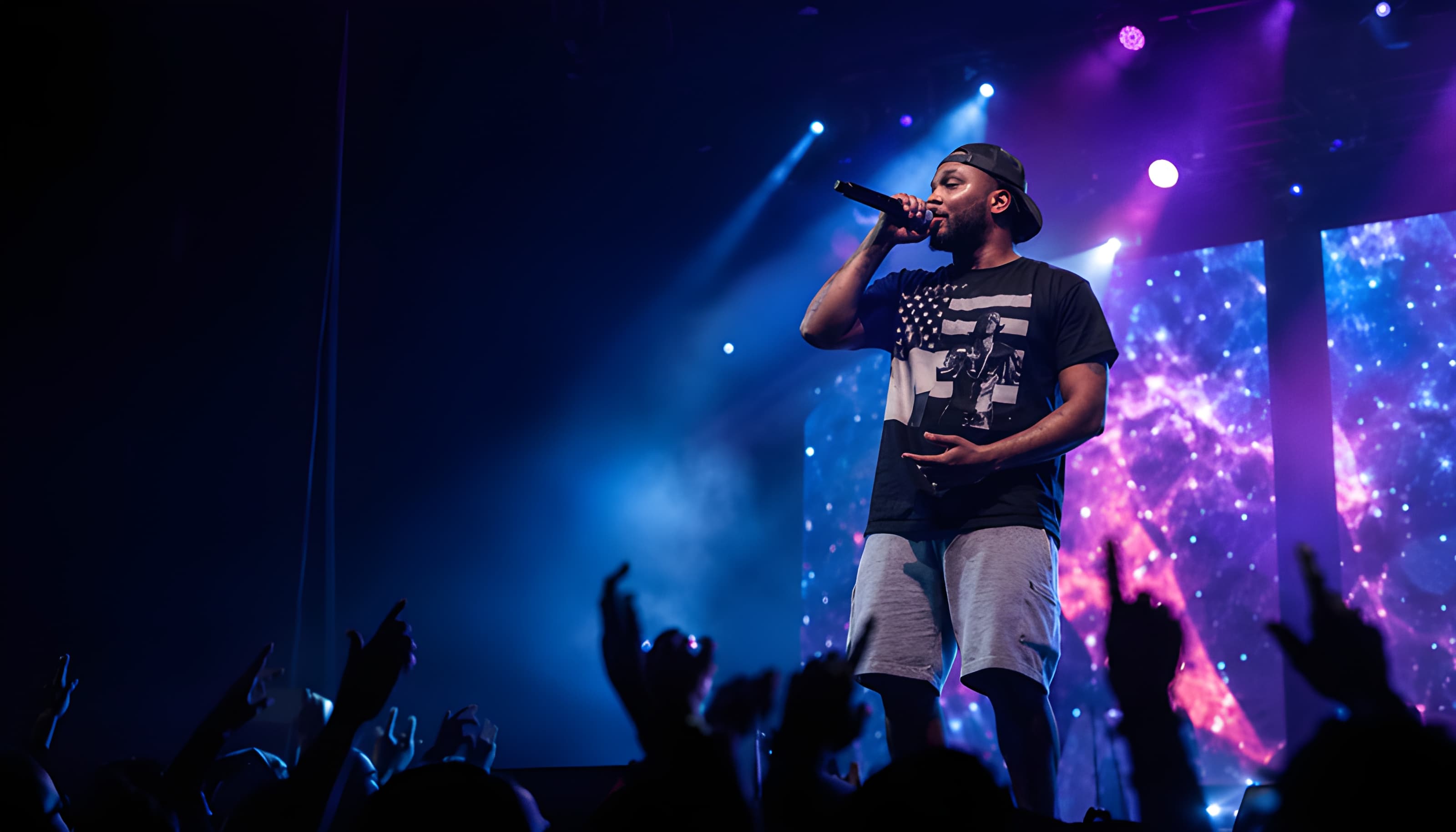 A male artist performs on stage under vibrant blue and purple lights for a silhouetted crowd.